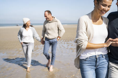 Lächelnde Frau hält einen Mann an der Hand, während sie mit einem Paar im Hintergrund am Strand spazieren geht - UUF21694