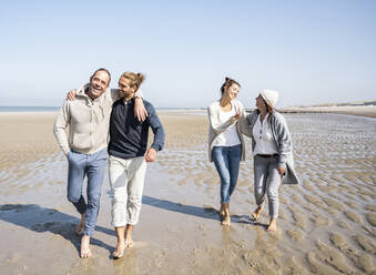 Smiling men waking with women talking in background at beach - UUF21710