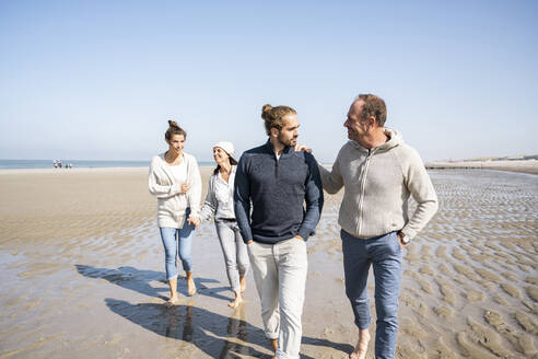 Vater und Schwiegersohn gehen mit einer lächelnden Frau im Hintergrund am Strand spazieren - UUF21713