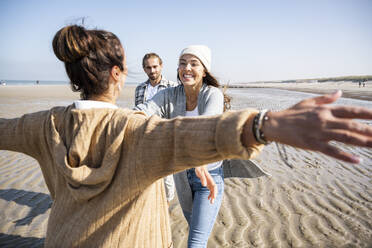 Woman with arms outstretched to hug daughter with men in standing in background at beach - UUF21727