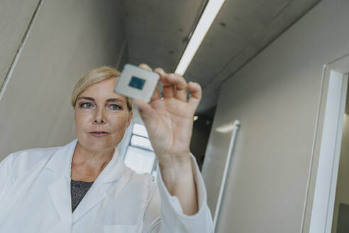 Blond scientist holding computer chip while standing at clinic corridor - MFF06337