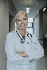 Smiling doctor with arms crossed standing at clinic corridor - MFF06341