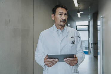 Scientist using digital tablet while standing at clinic corridor - MFF06356