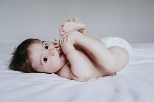 Niedlicher kleiner Junge, der den Fuß in den Mund nimmt, während er zu Hause auf dem Bett liegt - EBBF00874