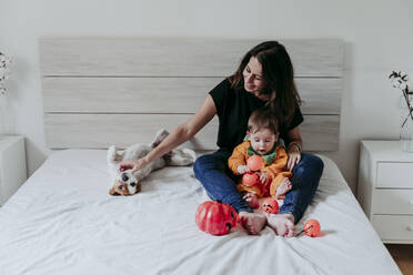 Mutter sitzt mit Sohn, der ein Halloween-Kostüm trägt, und Hund, der zu Hause auf dem Bett liegt - EBBF00876