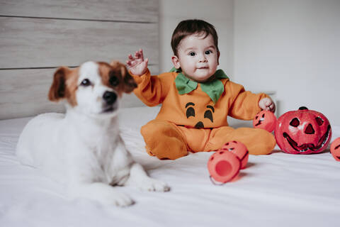 Baby-Junge trägt Halloween-Kostüm sitzt mit Hund auf dem Bett zu Hause, lizenzfreies Stockfoto