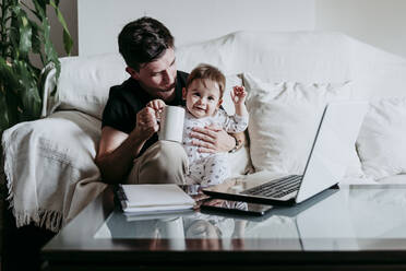 Father holding coffee cup with son sitting on sofa at home - EBBF00882