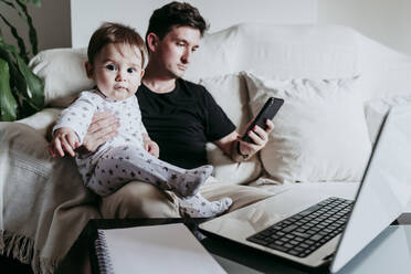 Father using smart phone while holding son sitting on sofa at home - EBBF00884