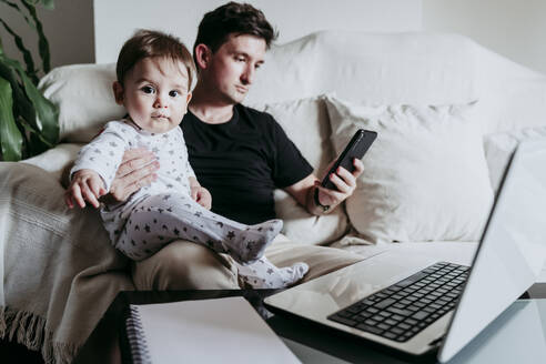 Vater benutzt Smartphone, während er seinen Sohn auf dem Sofa zu Hause hält - EBBF00884
