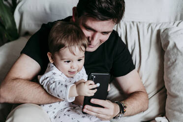 Vater und Sohn benutzen ein Smartphone, während sie zu Hause auf dem Sofa sitzen - EBBF00885
