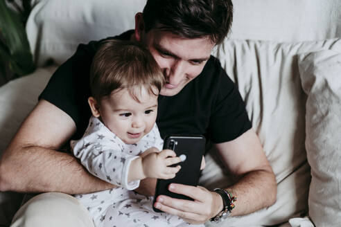 Vater und Sohn benutzen ein Smartphone, während sie zu Hause auf dem Sofa sitzen - EBBF00885