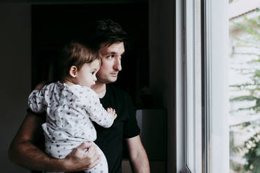 Father carrying son while standing looking through window at home - EBBF00887