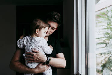 Vater hält seinen Sohn, während er durch das Fenster seines Hauses schaut - EBBF00888