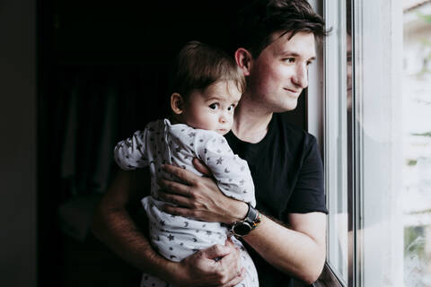 Lächelnder Vater, der seinen Sohn trägt, während er zu Hause durch das Fenster schaut, lizenzfreies Stockfoto