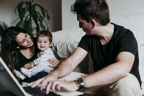 Lächelnde Mutter hält ihren Sohn, während der Vater zu Hause am Laptop arbeitet - EBBF00891
