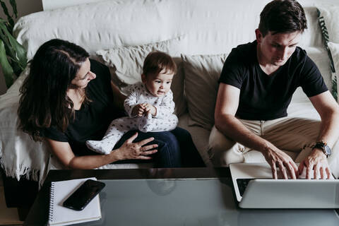 Sohn sitzt auf dem Schoß der Mutter und schaut dem Vater zu, der zu Hause am Laptop arbeitet, lizenzfreies Stockfoto