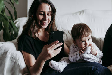 Mother text messaging on smart phone with son sitting on sofa at home - EBBF00894