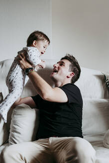 Fröhlicher Vater, der seinen Sohn abholt, während er zu Hause auf dem Sofa sitzt - EBBF00897