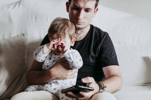 Vater benutzt sein Smartphone, während er mit seinem Sohn zu Hause auf dem Sofa sitzt - EBBF00900