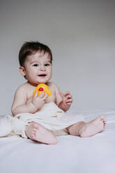 Cute boy playing with duck toy while sitting on bed at home - EBBF00902