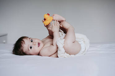 Cute baby boy playing with duck toy while lying down on bed at home - EBBF00904