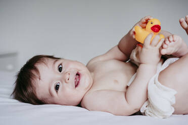 Smiling cute baby boy playing with duck toy while lying down on bed at home - EBBF00905