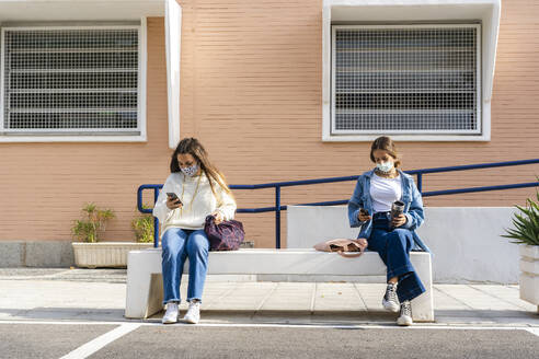 Female teenage friends social distancing while using smart phone sitting on concrete bench - ERRF04569