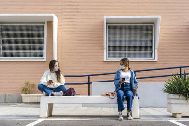 Freundinnen, die auf einer Betonbank sitzend eine Gesichtsmaske tragen und sich sozial distanzieren - ERRF04571