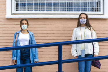 Female friends wearing protective face mask social distancing against wall - ERRF04578