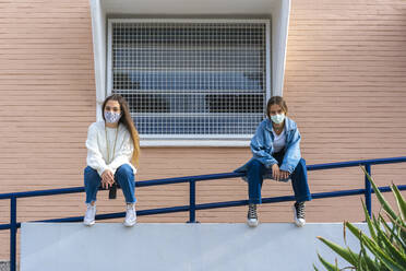 Teenage friends social distancing while wearing protective face mask sitting on railing during covid-19 - ERRF04582