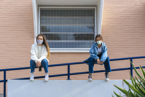 Teenage friends social distancing while wearing protective face mask sitting on railing during covid-19 - ERRF04582