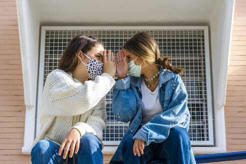 Female friends wearing protective mask sitting face to face on railing during sunny day - ERRF04587