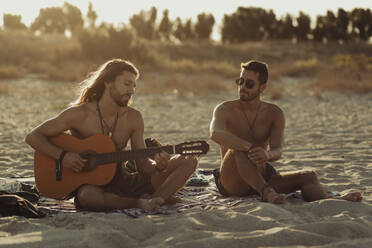 Entspannter talentierter Mann, der Akustikgitarre für einen männlichen Freund spielt, während er am Sandstrand bei Sonnenuntergang zusammensitzt - ADSF16686