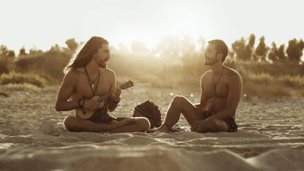Relaxed talented man playing acoustic guitar for male friend while sitting together on sandy beach at sunset - ADSF16691