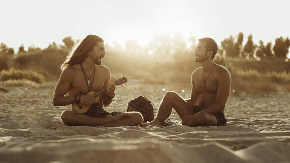 Entspannter talentierter Mann, der Akustikgitarre für einen männlichen Freund spielt, während er am Sandstrand bei Sonnenuntergang zusammensitzt - ADSF16691