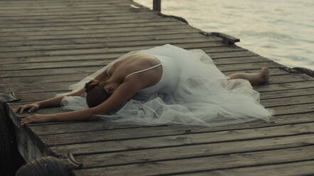 Unrecognizable elegant female ballet dancer in white tutu performing graceful seated pose on wooden pier near sea at sunset time - ADSF16692