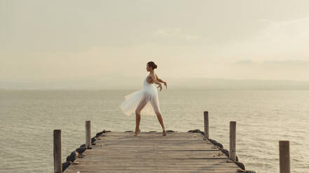Ganzkörper junge anmutige Tänzerin in weißem Kleid stehen auf Zehenspitzen bei der Durchführung von Ballett-Bewegungen auf hölzernen Promenade gegen Meer im Sommer Abend - ADSF16693