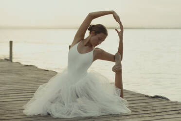 Full body flexible classic ballet female dancer in elegant white dress performing sensual pose on wooden pier against blurred sea in summer evening - ADSF16694