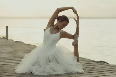 Ganzkörper flexible klassische Ballett Tänzerin in eleganten weißen Kleid Durchführung sinnliche Pose auf hölzernen Pier gegen verschwommene Meer im Sommer Abend - ADSF16694