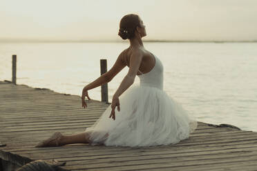 Full body flexible classic ballet female dancer in elegant white dress performing sensual pose on wooden pier against blurred sea in summer evening - ADSF16695