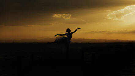 Silhouette einer eleganten, biegsamen Tänzerin mit ausgestreckten Armen, die vor einem wolkenverhangenen Himmel Ballettbewegungen ausführt - ADSF16696