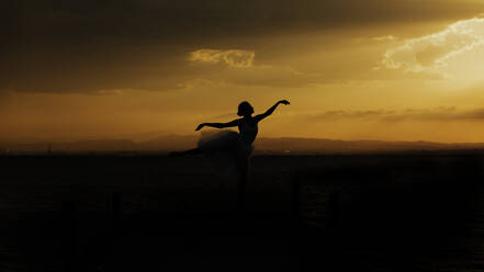 Silhouette of elegant flexible female dancer with outstretched arms performing ballet movement against cloudy sundown sky - ADSF16696