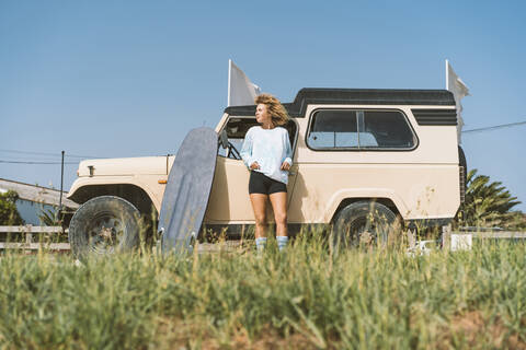 Young Afro woman looking away while standing with surfboard against old off-road vehicle on sunny day stock photo