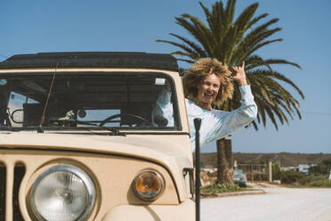 Fröhliche blonde Frau gestikuliert, während sie in einem alten Geländewagen an einem sonnigen Tag sitzt - DAMF00573