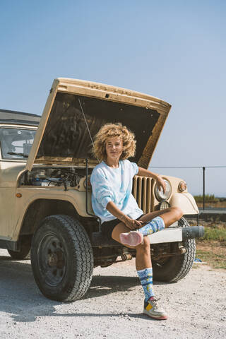 Confident young blond Afro woman looking away while sitting on bumper of broken-down vehicle during sunny day stock photo