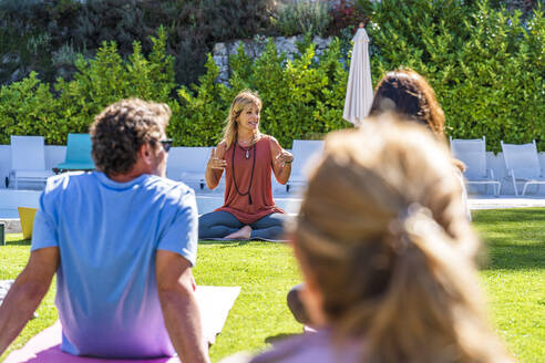 Blonde Yogalehrerin unterrichtet Touristen beim Sitzen im Freien an einem sonnigen Tag - DLTSF01359