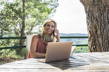 Glückliche Frau, die über ihr Smartphone spricht, während sie mit ihrem Laptop an einem Tisch im Gesundheitszentrum an einem sonnigen Tag sitzt - DLTSF01378