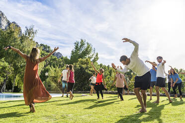 Reife weibliche Fitness-Instruktorin tanzt mit Touristen bei Gesundheit Rückzug auf Gras gegen Himmel während des sonnigen Tages - DLTSF01387