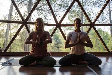 Männliche und weibliche Yogalehrer beim Üben des Lotussitzes vor einem Fenster in einem Gesundheitszentrum - DLTSF01398