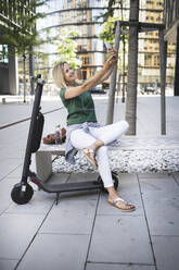 Mid erwachsene Frau hört Musik und nimmt selfie beim Sitzen auf Bank in der Stadt - HMEF01109